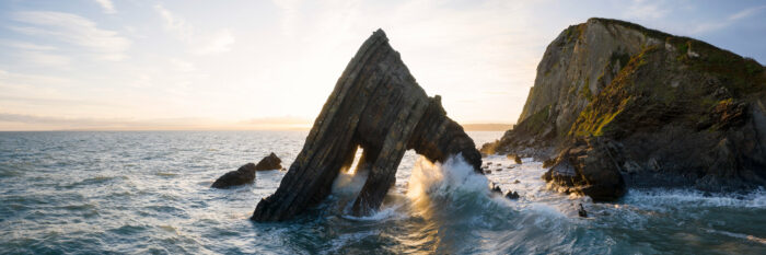 A panoramic print as a wave crashes against rocky coastline